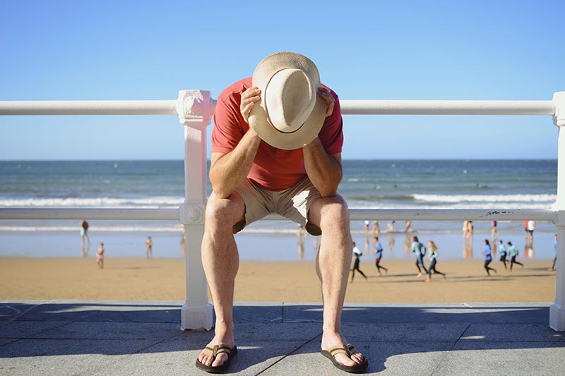 Man clasping head sitting on a rail at beach.